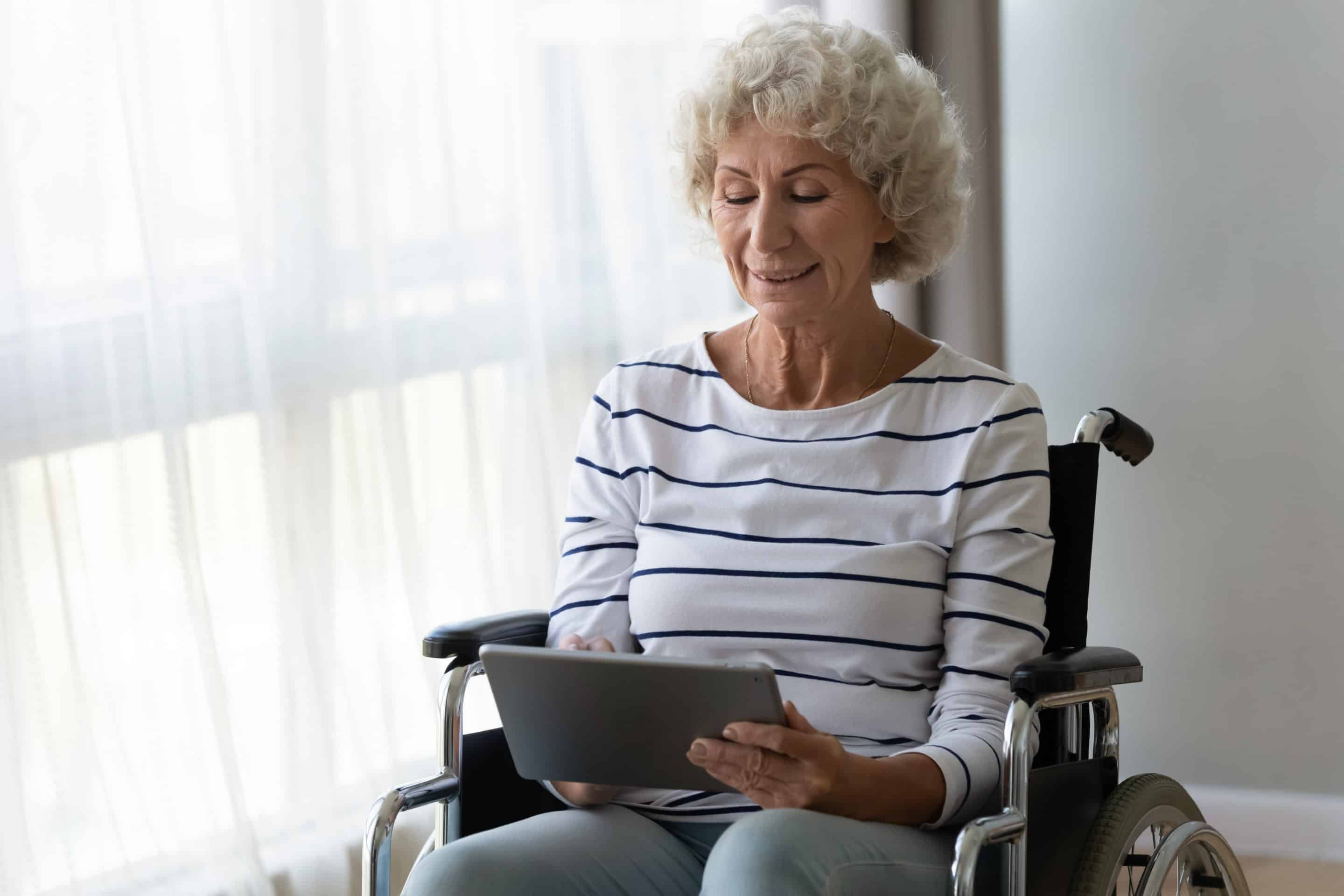 An older woman in a wheelchair smiling while using a tablet by a sunlit window